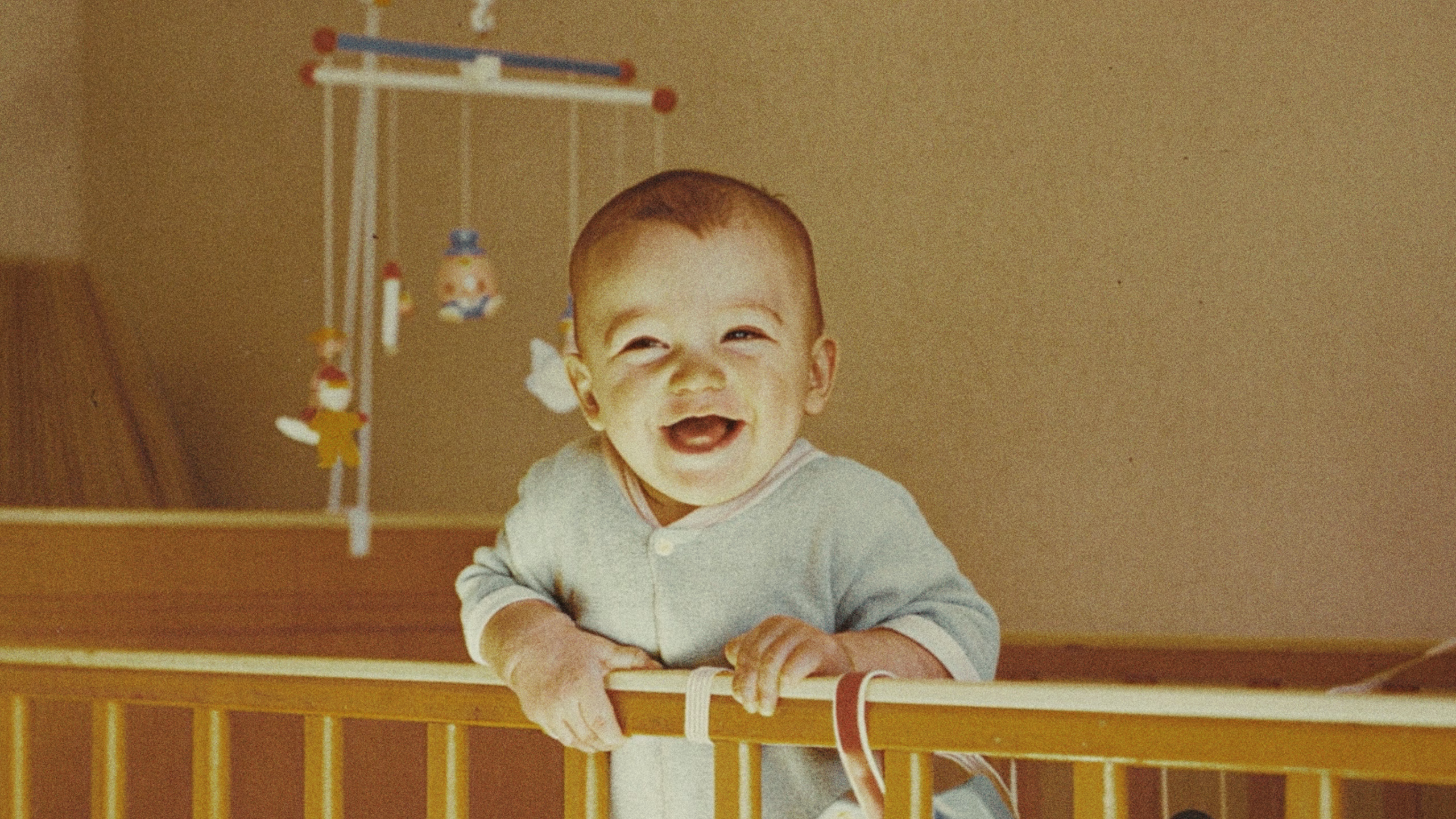 BabyChadwell » Tree9 Films Smiling baby leaning over the railing of a wooden crib, wearing a light blue onesie.